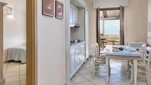 Una cocina con armarios blancos, una mesa de comedor con sillas y vista a la playa a través de una puerta abierta.