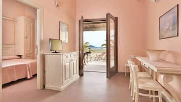 A room with a bed, a desk with chairs, a cabinet, and a view of the beach through an open door.