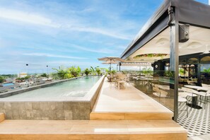 Un área de piscina en la azotea con vistas a la ciudad, mobiliario exterior y una zona de bar techada.
