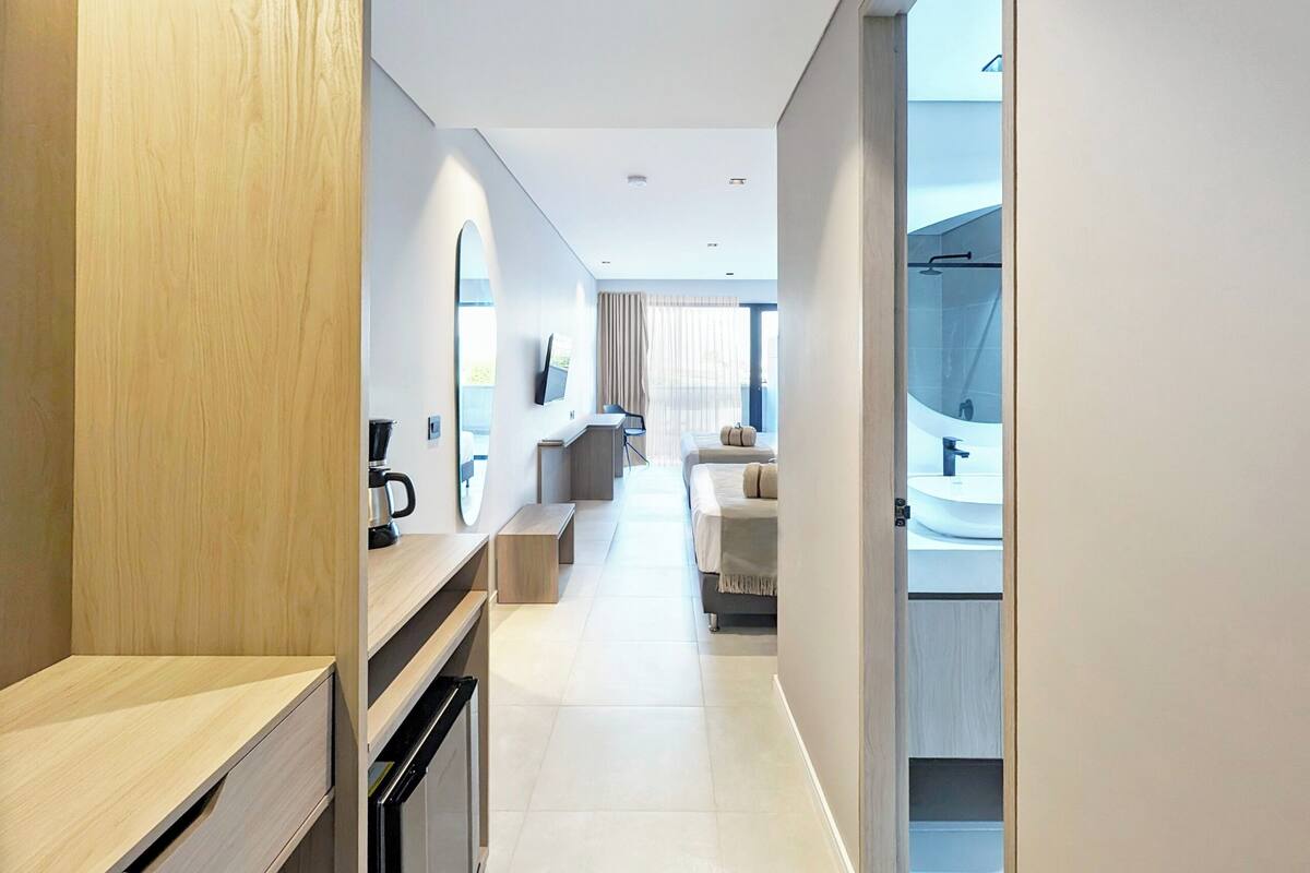 A modern hotel room with a wooden desk, a bed, a sofa, a small kitchen area, and a bathroom with a large mirror.