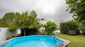 Una piscina rectangular con plataforma de madera, rodeada de vegetación y un molino de viento al fondo.