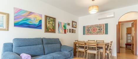 A living room with a blue sofa, wooden dining table, and chairs, with various framed artworks on the walls.