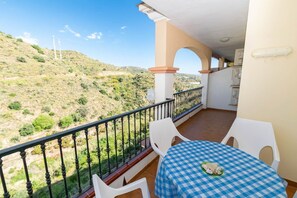 A balcony with a checkered tablecloth, white chairs, and a view of a hilly landscape.