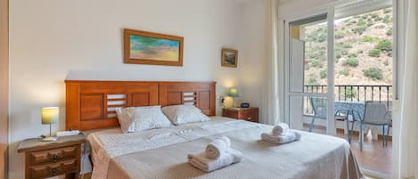 A hotel room with a bed, a nightstand, a lamp, a painting, a window with a view, and a balcony with chairs.