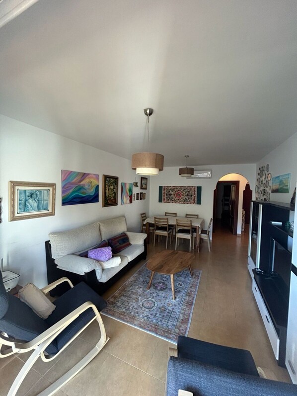 A living room with a sofa, coffee table, and dining area. There are paintings on the walls, a TV, and a fireplace.