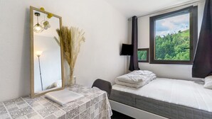 Een slaapkamer met een bed, een bureau met een laptop, een spiegel, een raam met uitzicht op groen en een tv aan de muur.