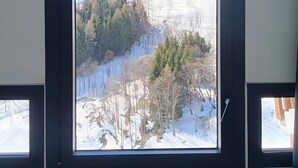 Een kamer met een groot raam, met uitzicht op een besneeuwd landschap met bomen en een heuvel.