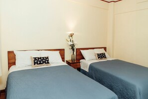 Habitación de hotel con dos camas individuales, cada una con una colcha azul y un almohadón blanco con un diseño geométrico negro. Hay una mesita de noche entre las camas con un florero.