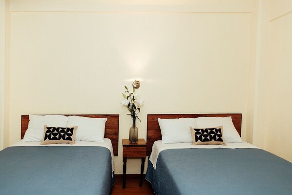 Habitación de hotel con dos camas, cada una con una colcha azul y una almohada de estampado geométrico. Una pequeña mesita de noche con un florero se encuentra entre las camas.