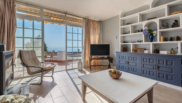 Un salon comprenant un téléviseur, une table, un canapé et un fauteuil. On y trouve également des étagères remplies de divers objets, et la vue donne sur la mer.