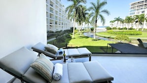 A balcony with sun loungers, a small table with a bottle and glass, and a view of a well-maintained lawn with palm trees and a building in the background. - Beachfront condo in the heart of Nuevo Vallarta (Nuevo Vallarta)