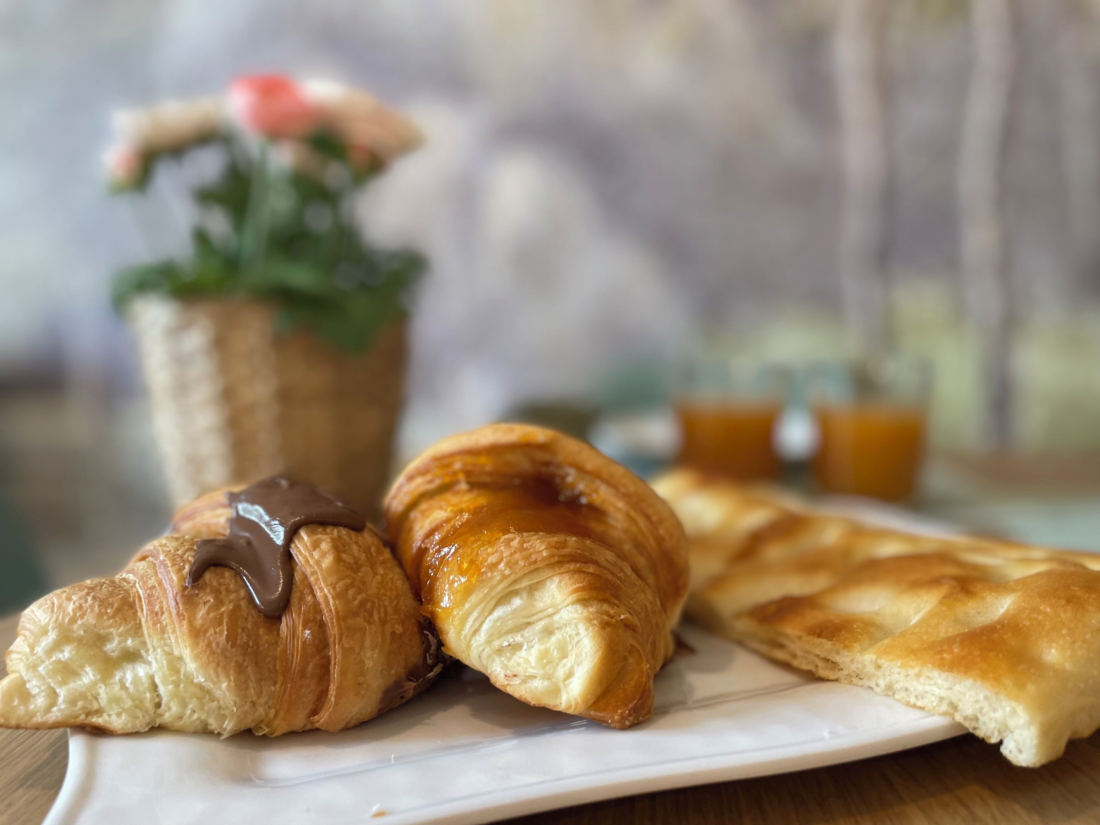 A plate with two croissants and a piece of bread, a small basket with a plant, and a cup of tea on a wooden table.