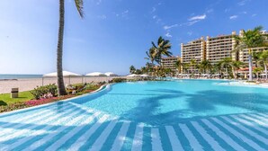 A resort pool with clear blue water, surrounded by palm trees and lounge chairs. - Beachfront condo best location spectacular views (Puerto Vallarta)