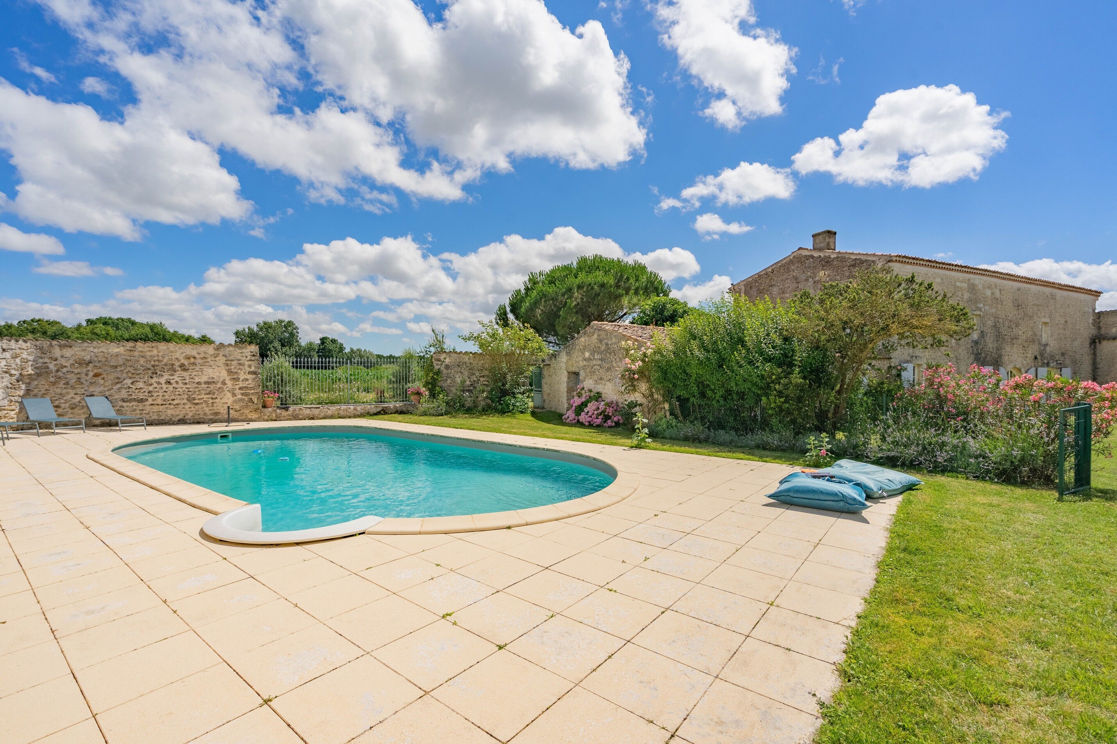 A swimming pool with a curved edge, surrounded by a tiled patio, a stone wall, and a building with a tiled roof.