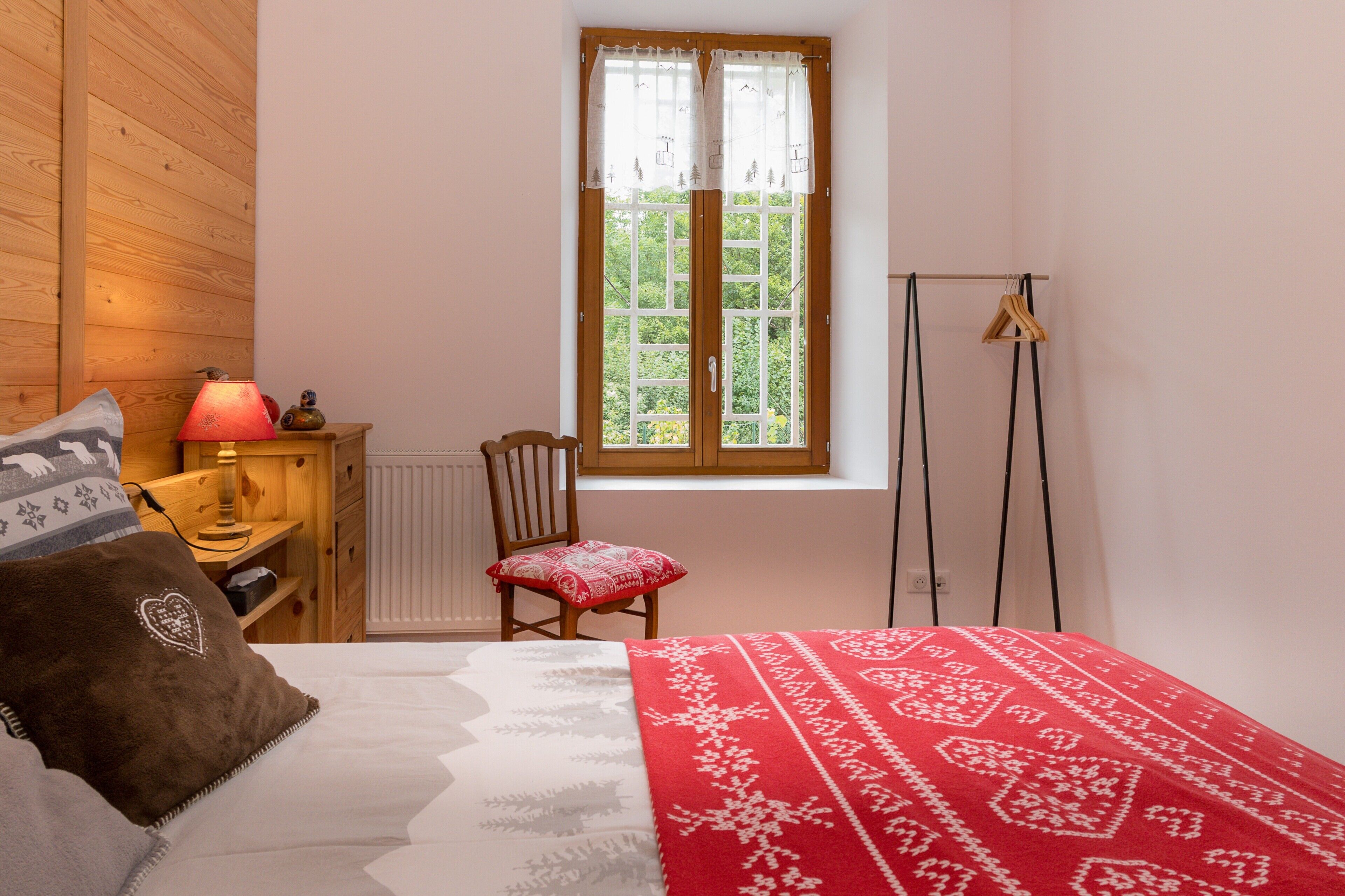 A bedroom with a wooden floor, a window with curtains, a chair, a bedside table with a lamp, and a bed with a patterned blanket.