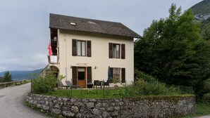A two-story house with a stone wall, a wooden door, and a small porch with outdoor furniture. - La Sabaudia En Chartreuse Apartment with Mountain View, Private Terrace, and Wi-Fi (Saint-Pierre-d'Entremont)