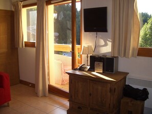 A room with a sliding glass door leading to a balcony, a television mounted on the wall, a wooden cabinet, and a desk with a lamp.