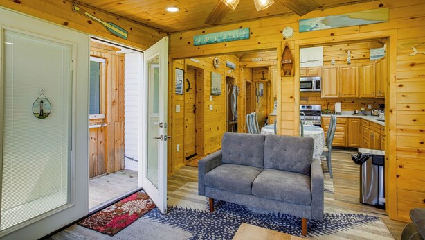 A cozy cabin interior with a living room featuring a gray sofa, wooden walls, a ceiling fan, and a kitchen area with wooden cabinets and appliances.
