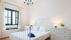Una cama de metal blanco con cabecera, una mesita de noche con lámpara y una ventana con cortinas.