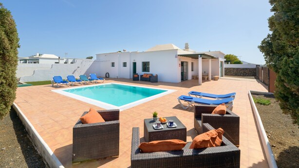 Un área de piscina con sillones, una mesa de comedor con una bebida, y un edificio blanco al fondo.