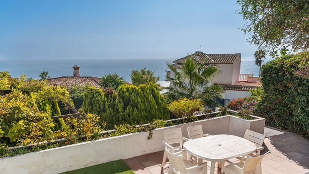 Eine Terrasse mit Tisch und Stühlen, Blick aufs Meer, und ein Gebäude mit Ziegeldach.