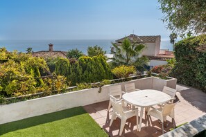 Eine Terrasse mit Tisch und Stühlen, Blick aufs Meer, und ein Gebäude mit Ziegeldach.
