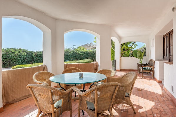 Un patio cubierto con mobiliario de mimbre, una mesa de vidrio y vistas a zonas verdes y un edificio.