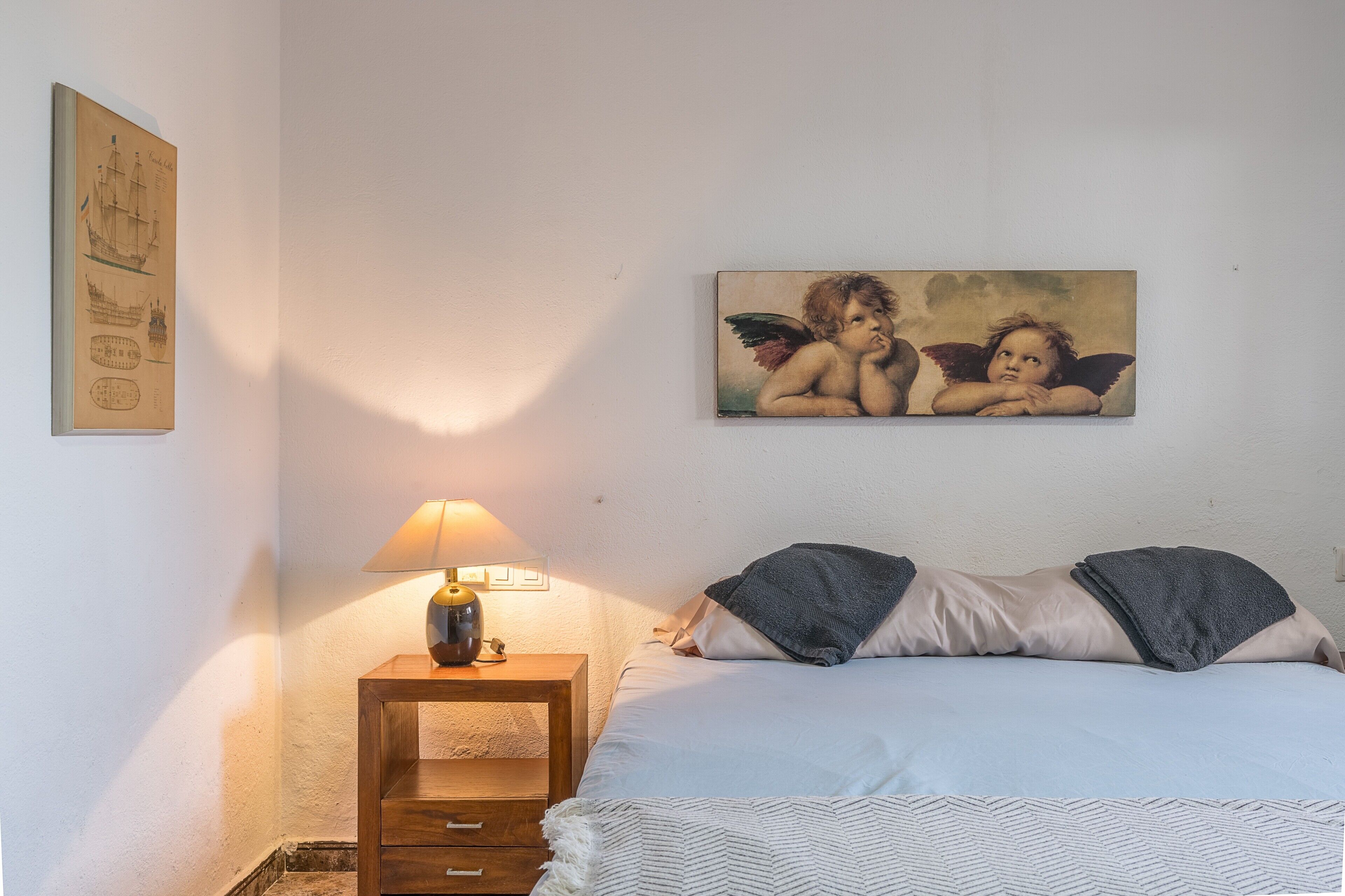 A bedroom with a bed, a nightstand with a lamp, and a painting of cherubs on the wall.