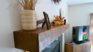 Une cheminée surmontée d’une étagère où sont disposés un vase contenant des plantes séchées, un cadre décoratif et un morceau de bois flotté. Un téléviseur est fixé sur un meuble en bois à l’arrière-plan.