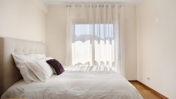 Une chambre à coucher avec un lit, une table de chevet et une fenêtre ornée de rideaux légers.