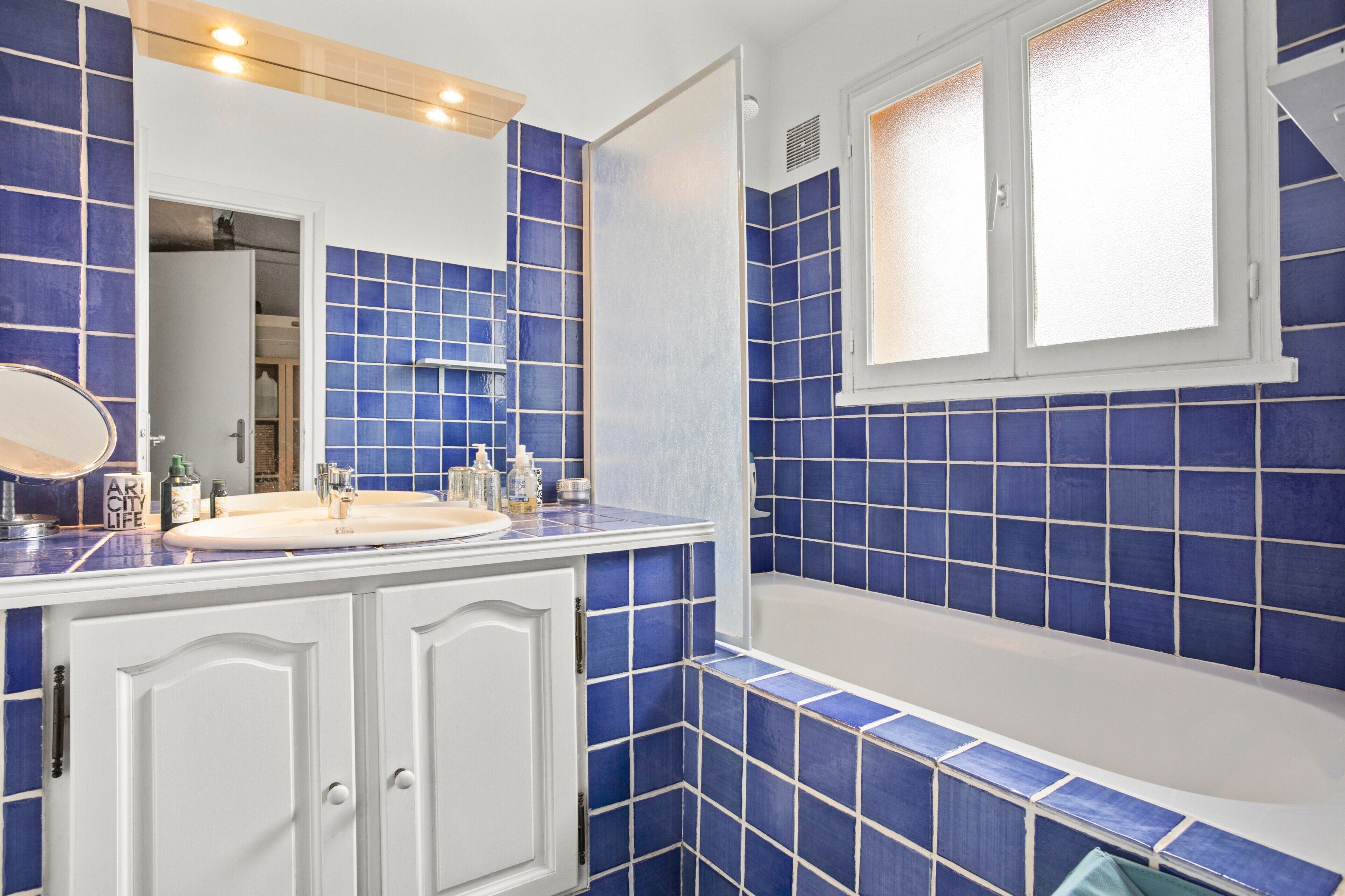 Une salle de bain avec des carreaux bleus, un lavabo blanc et une fenêtre au-dessus de la baignoire.