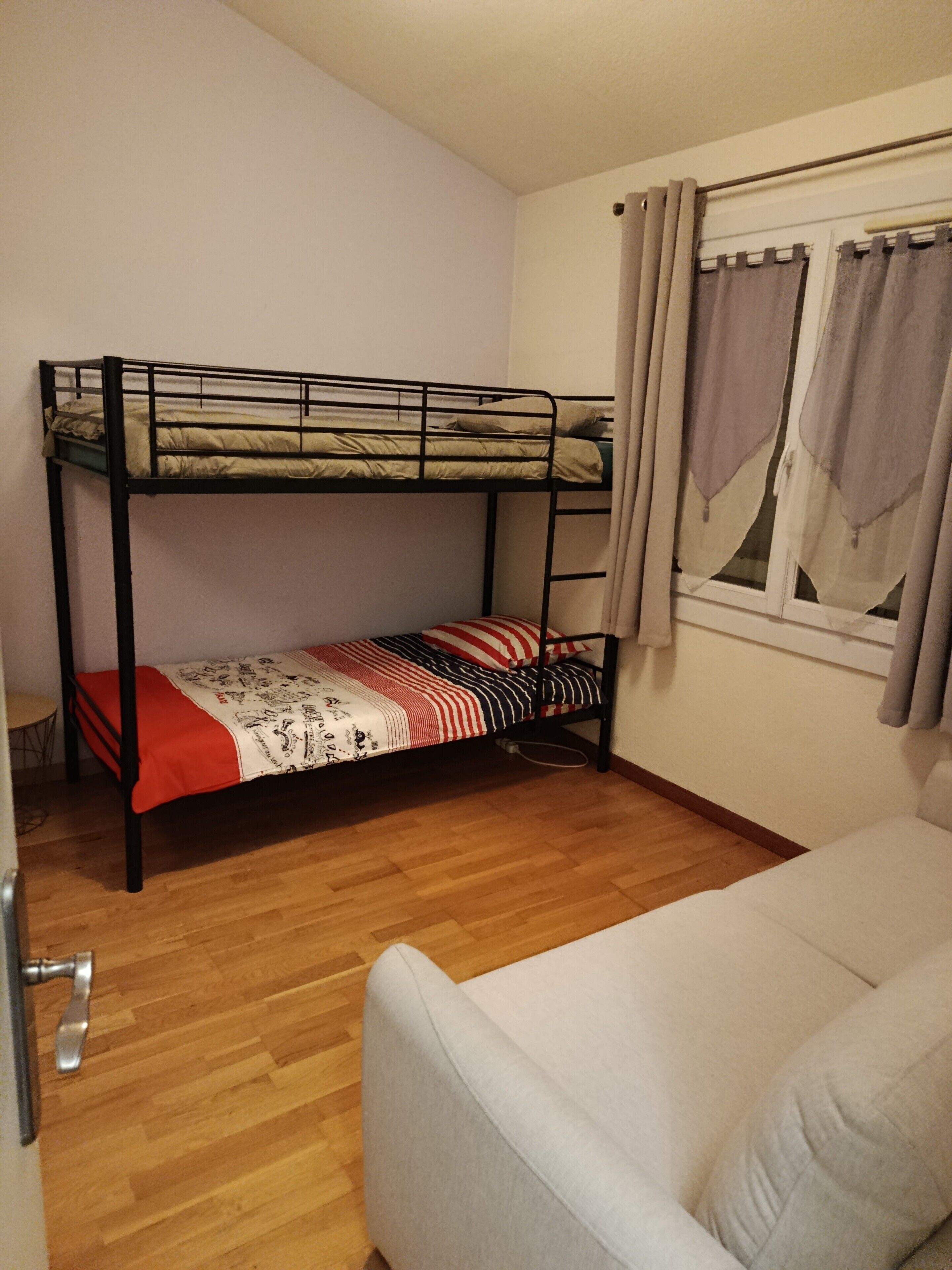 Ein Zimmer mit einem Etagenbett, einer Schlafcouch, einem Fenster mit Vorhängen und Holzboden.