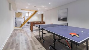 Une table de ping-pong dans un salon moderne, avec un escalier en bois et une cuisine en arrière-plan.