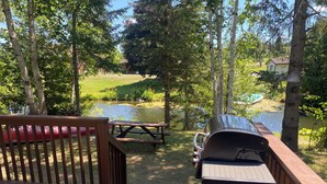 Une terrasse en bois avec un barbecue et une table de pique-nique, donnant sur un étang et la verdure environnante.
