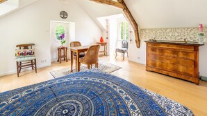 Ein geräumiges Dachzimmer mit freiliegenden Holzbalken, einem großen blauen Teppich mit Muster, einer Holzkommode und einem Essbereich mit Stühlen und Tisch.