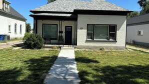Une maison à deux étages avec un toit gris, une porte d’entrée noire et un petit jardin devant, avec un chemin menant à l’entrée.