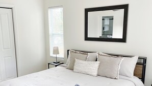 Una cama bien hecha con edredón blanco y toallas dobladas, un ventilador de techo, una mesita de noche con lámpara y un espejo grande en la pared.