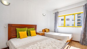 A hotel room with two beds, a wooden headboard, a bedside table, a window with yellow frames, and a view of buildings.