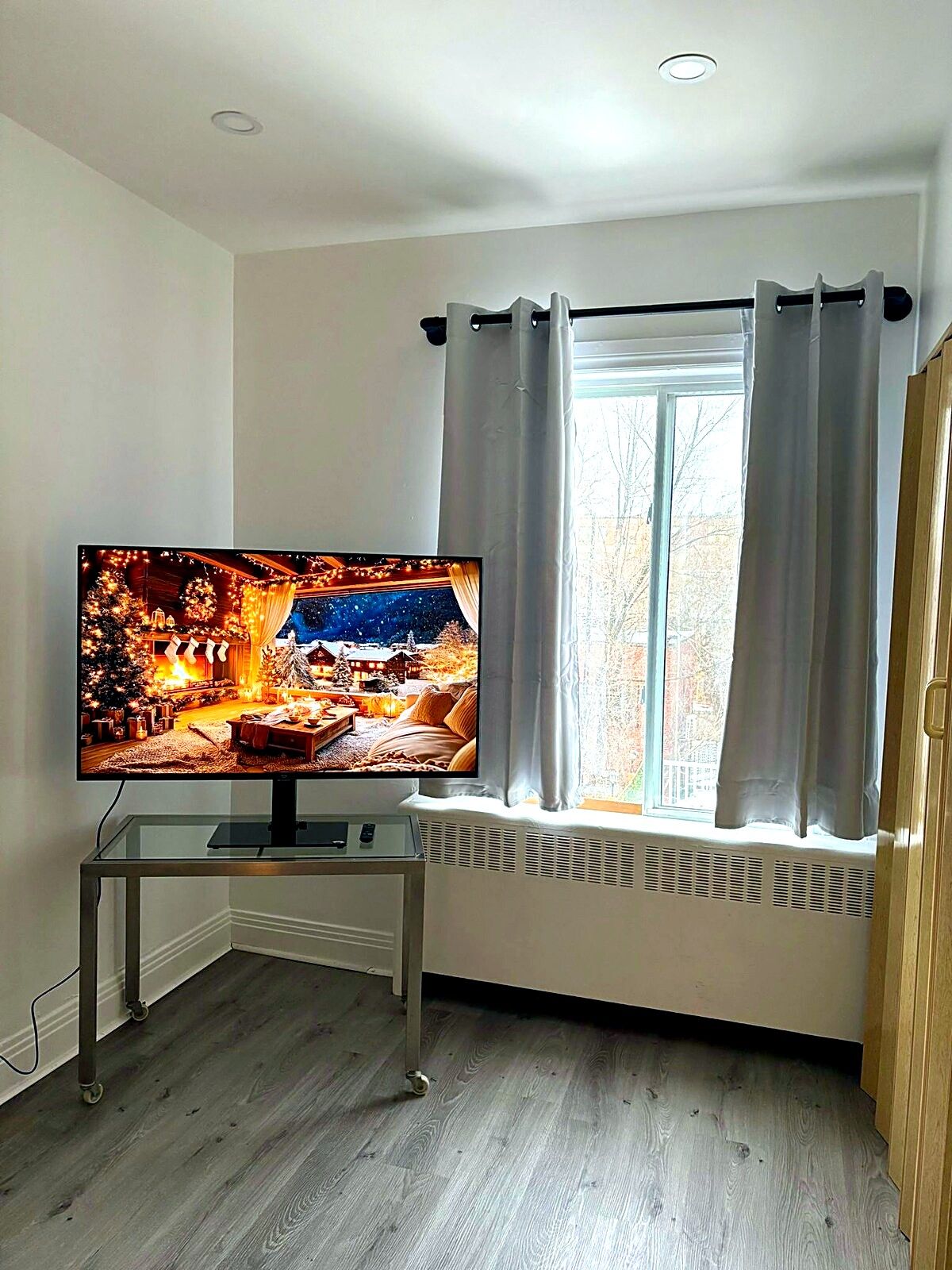 Quarto com televisão sobre suporte de vidro, janela com cortinas e radiador sob a janela.