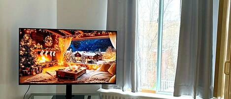 Huoneessa on televisio lasijalustalla, ikkunassa on verhot ja ikkunan alla on patteri.