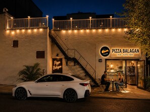 Une voiture blanche est garée devant un immeuble. On peut voir une enseigne indiquant « Pizza Balagan » et un escalier menant à un balcon.