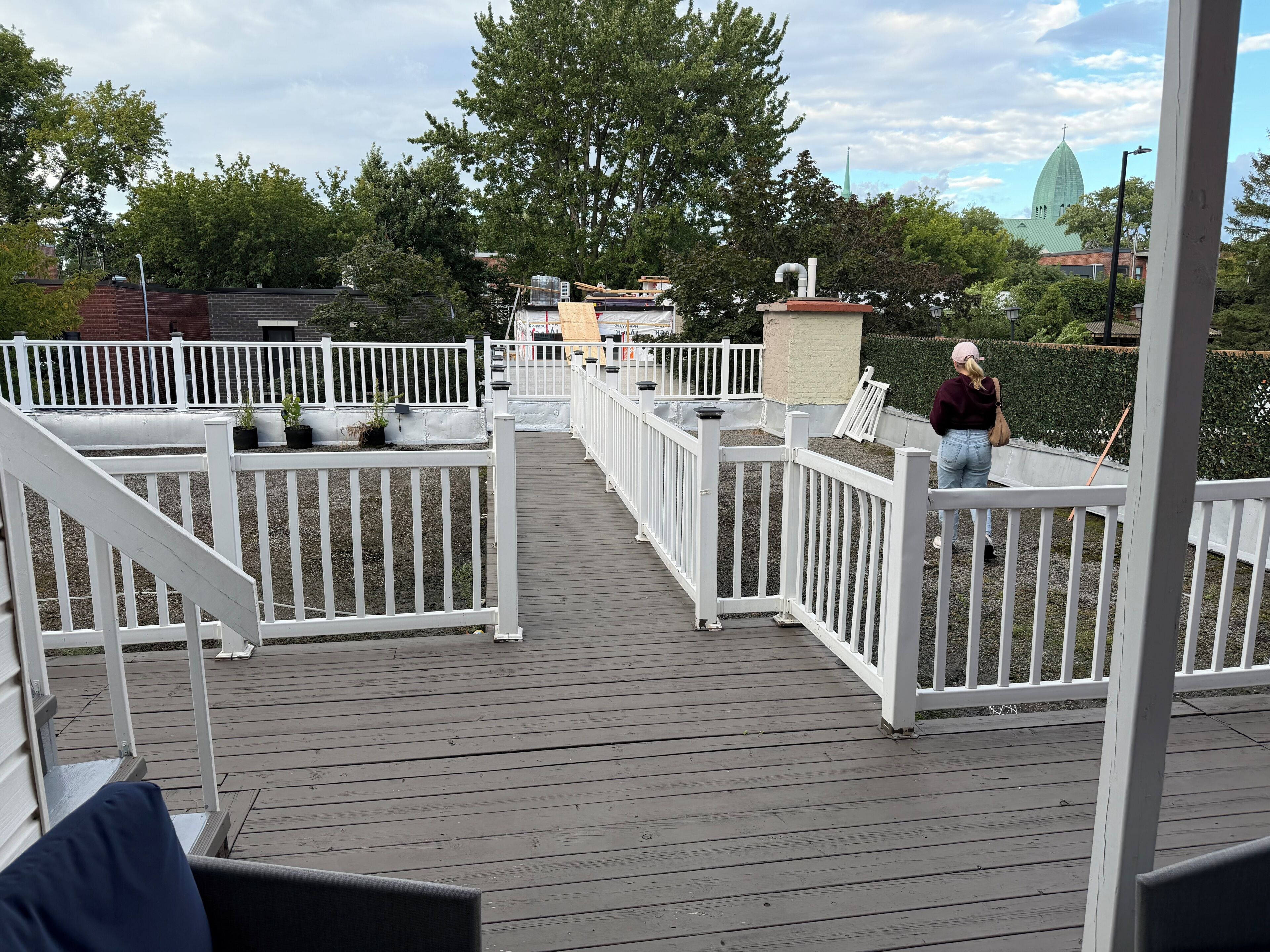 A wooden deck with white railings, potted plants, and a view of a church steeple in the background.