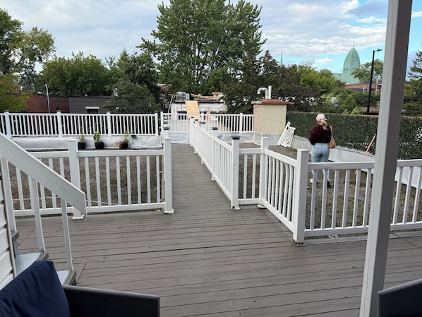 Une terrasse en bois avec des balustrades blanches, des plantes en pot et, au loin, la vue sur le clocher d’une église.
