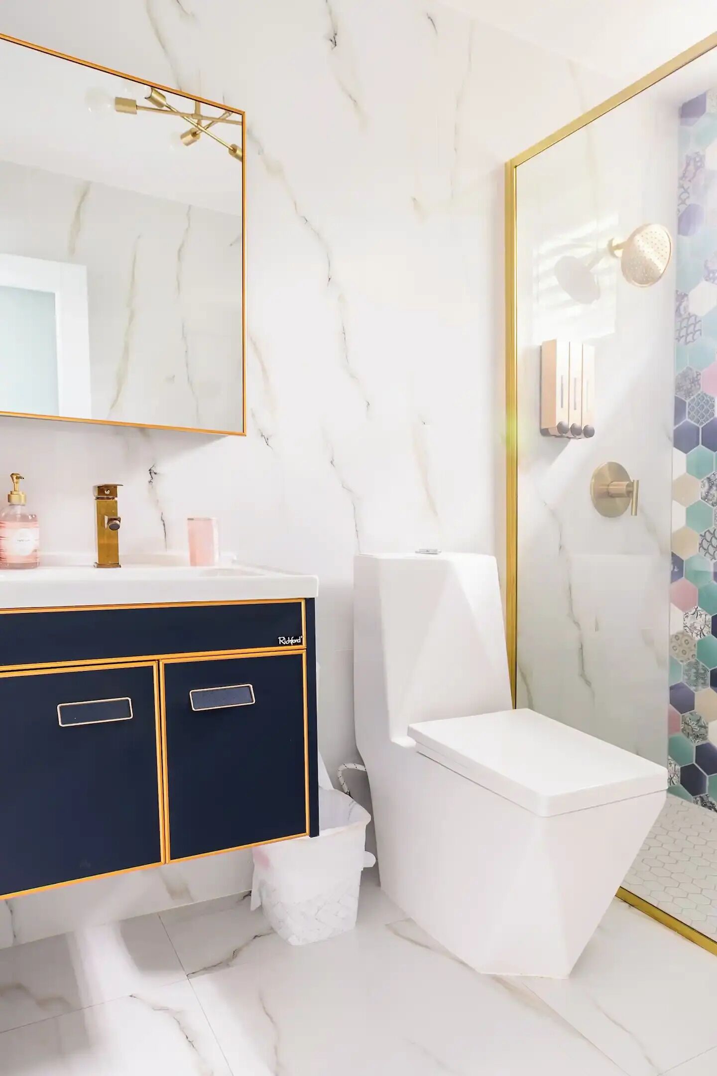 Un baño con pared de mármol blanco, espejo con marco dorado, tocador negro y dorado, inodoro blanco y ducha con cabezal dorado.