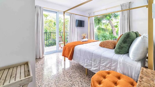 A bedroom with a bed, a TV, a balcony with curtains, and a wooden bench. - Siramada Village (Humacao)
