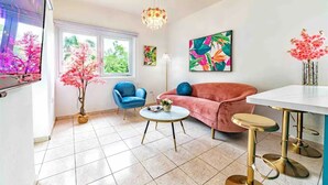 Sala de estar com sofá rosa, poltrona azul e mesa de centro redonda. Há uma janela com vista para árvores, um lustre e um quadro na parede.