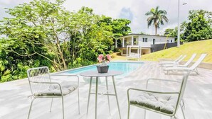 Uma área de piscina com espreguiçadeiras e uma mesa, rodeada por vegetação exuberante e uma casa ao fundo.