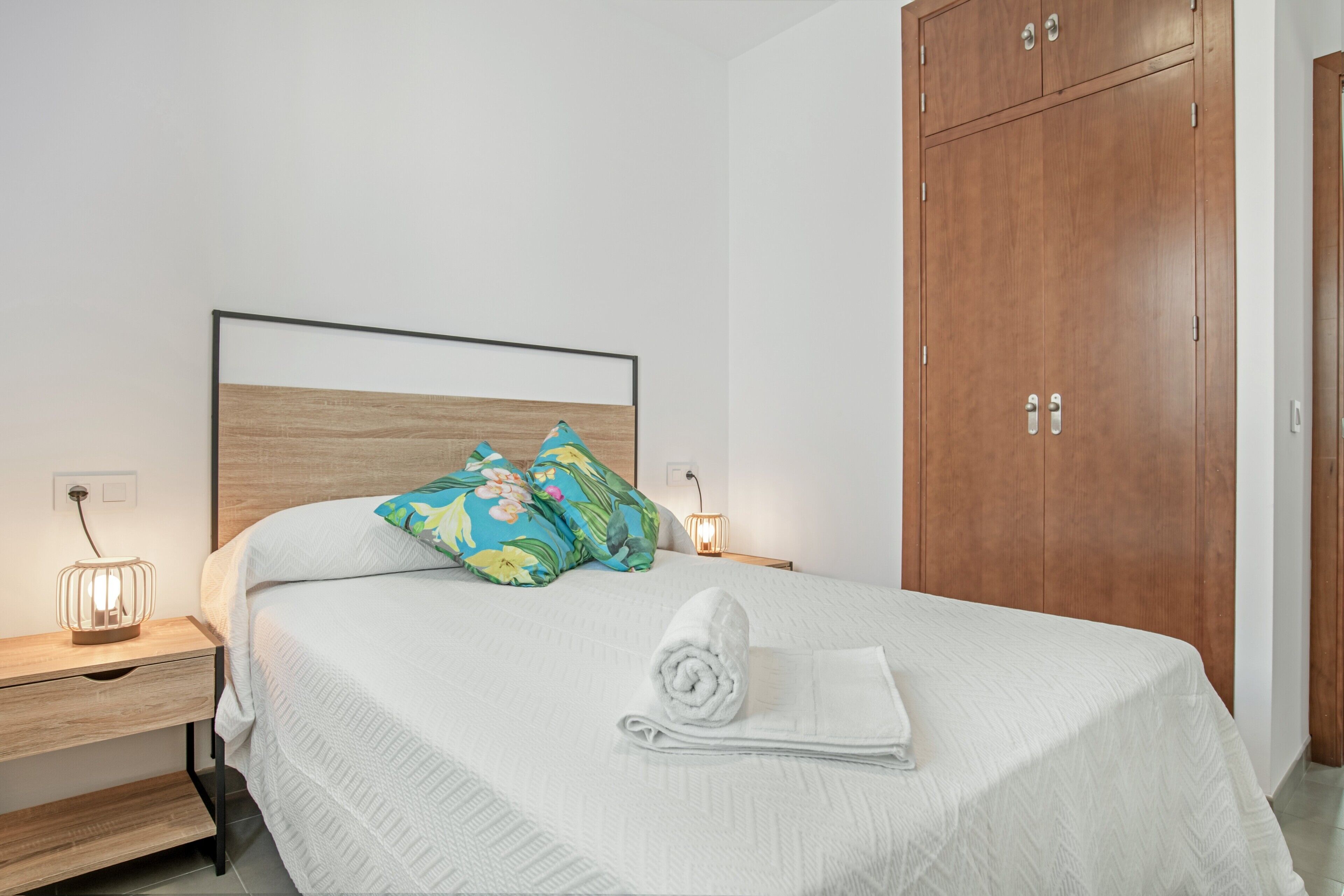 A neatly made bed with a white bedspread and a rolled towel, a wooden wardrobe, and a nightstand with a lamp.