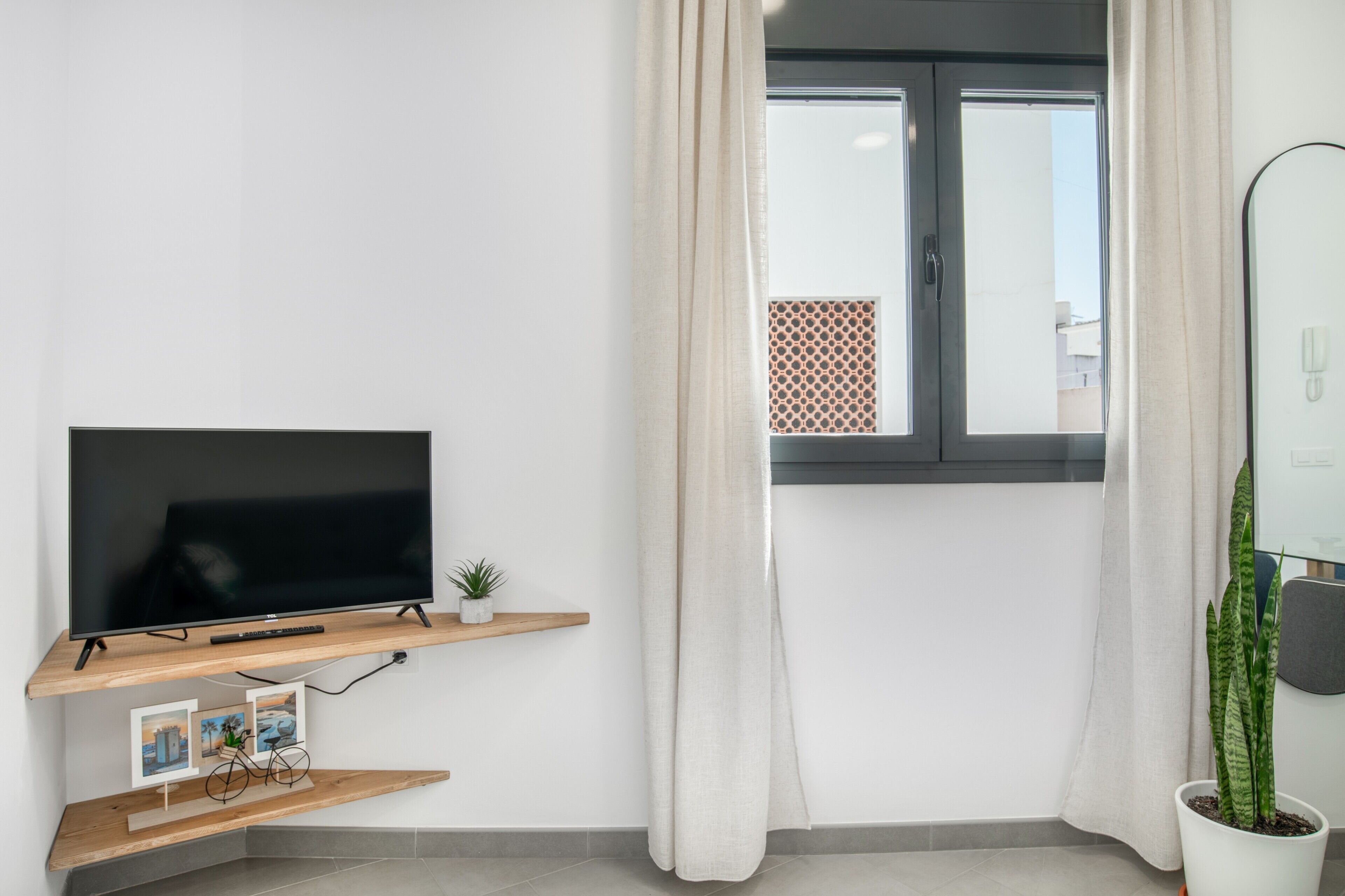 A modern room with a flat-screen TV on a wooden shelf, a potted plant, and a window with curtains.