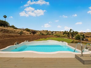 A swimming pool with a wooden deck, surrounded by a hilly landscape and clear blue sky.